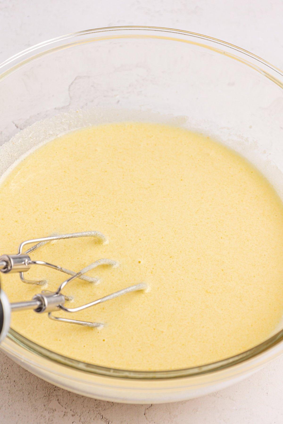 Un primo piano di una ciotola di vetro piena di impasto giallo chiaro. Due fruste metalliche di un frullino elettrico sono parzialmente visibili sul lato destro della ciotola. La ciotola è posta su una superficie chiara.