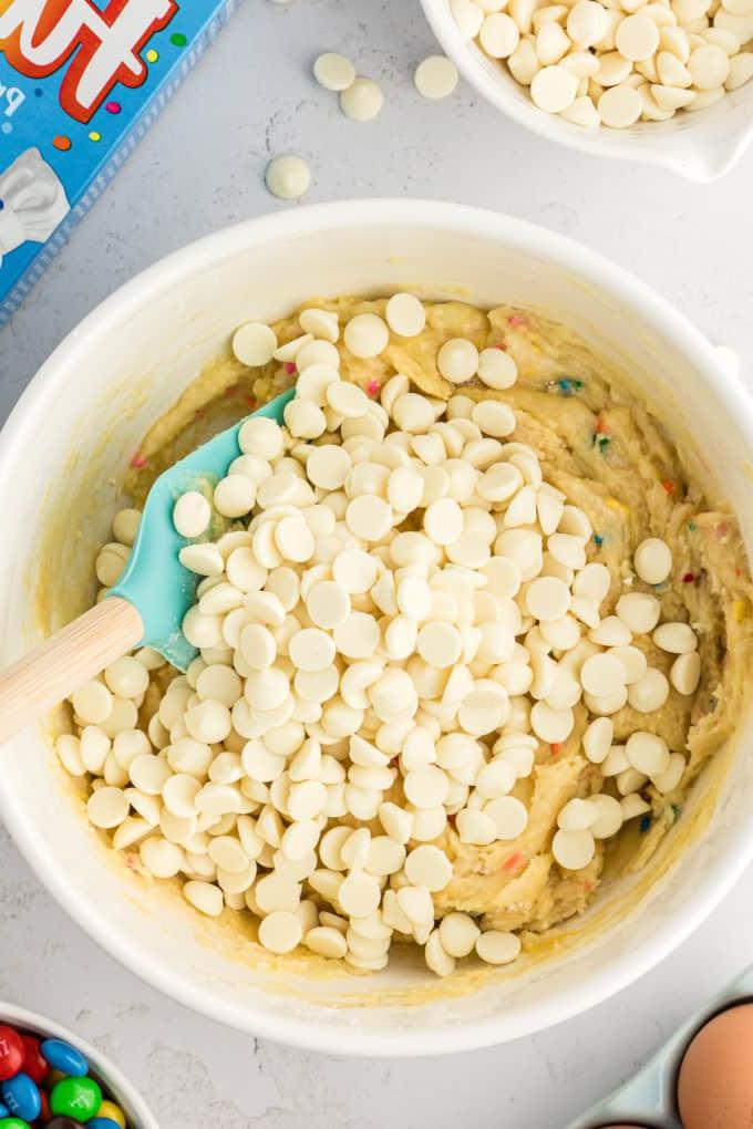 Barrette di biscotti da preparato per torte 10 a white bowl full of funfetti cake bar mix with a rubber spatula in it and a pile of white chocolate chips on top, a cake mix box, eggs, a bowl of M&Ms, and a bowl of white chocolate chips surrounding the bowl