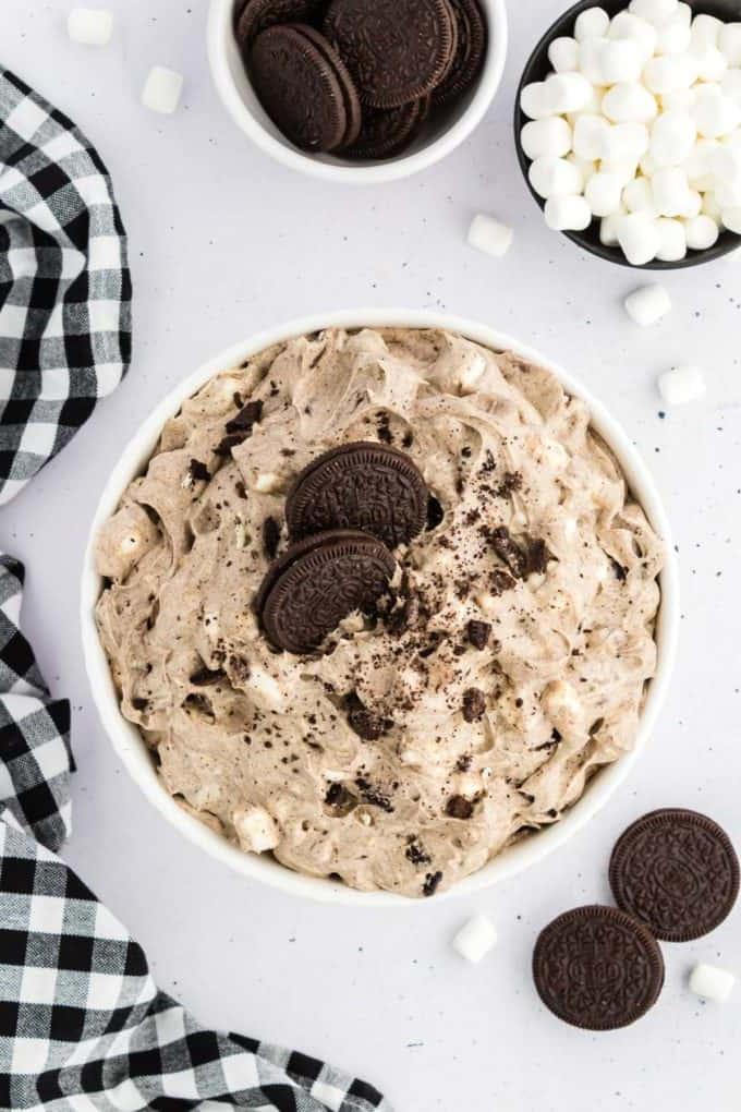 Ricetta Crema Soffice Oreo (Senza Formaggio Cremoso) 10 Fluff di Oreo sormontato da briciole di Oreo e due Oreo interi in una ciotola di vetro circondato da Oreo, mini marshmallow e un panno da cucina a scacchi bianchi e neri