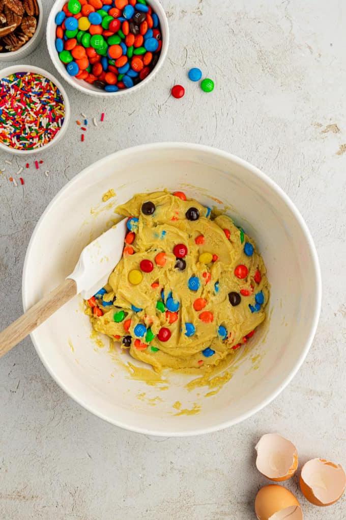 Fare Biscotti dal Mix per Torte 10 Impasto per biscotti con preparato per torte in una ciotola bianca con con M&M's mescolati con una spatola bianca e di legno circondata da una ciotola di codette, una ciotola di M&M's e una ciotola di noci