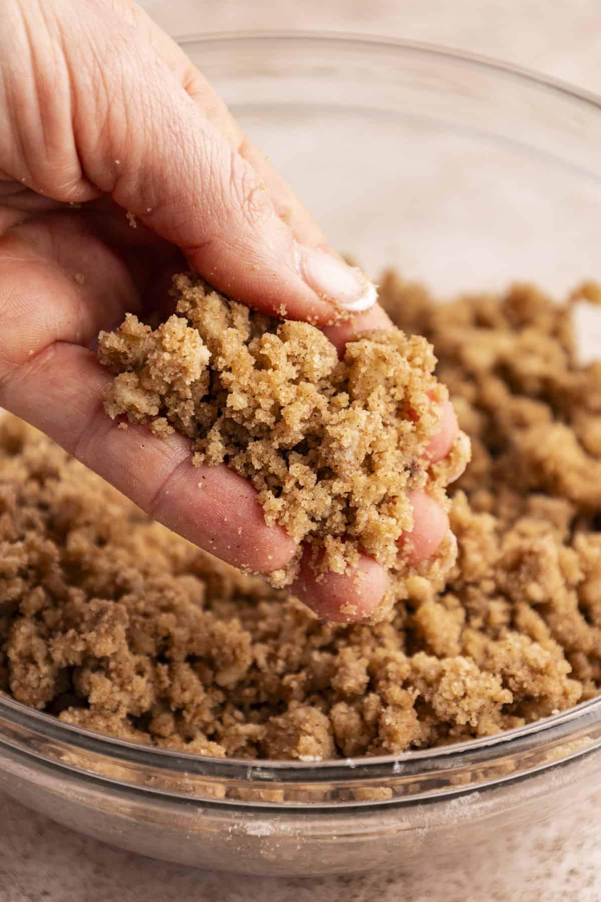 Migliore Ricetta per Budino di French Toast 10 Una mano che tiene un topping croccante sopra una ciotola di vetro trasparente piena di altro topping croccante.