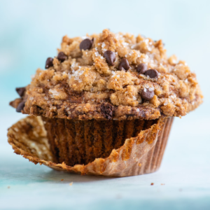 Ricetta della Copertura Sbriciolata 10 Un primo piano di un muffin con gocce di cioccolato e una dorata copertura streusel, parzialmente srotolato dal suo pirottino di carta marrone, su sfondo azzurro.