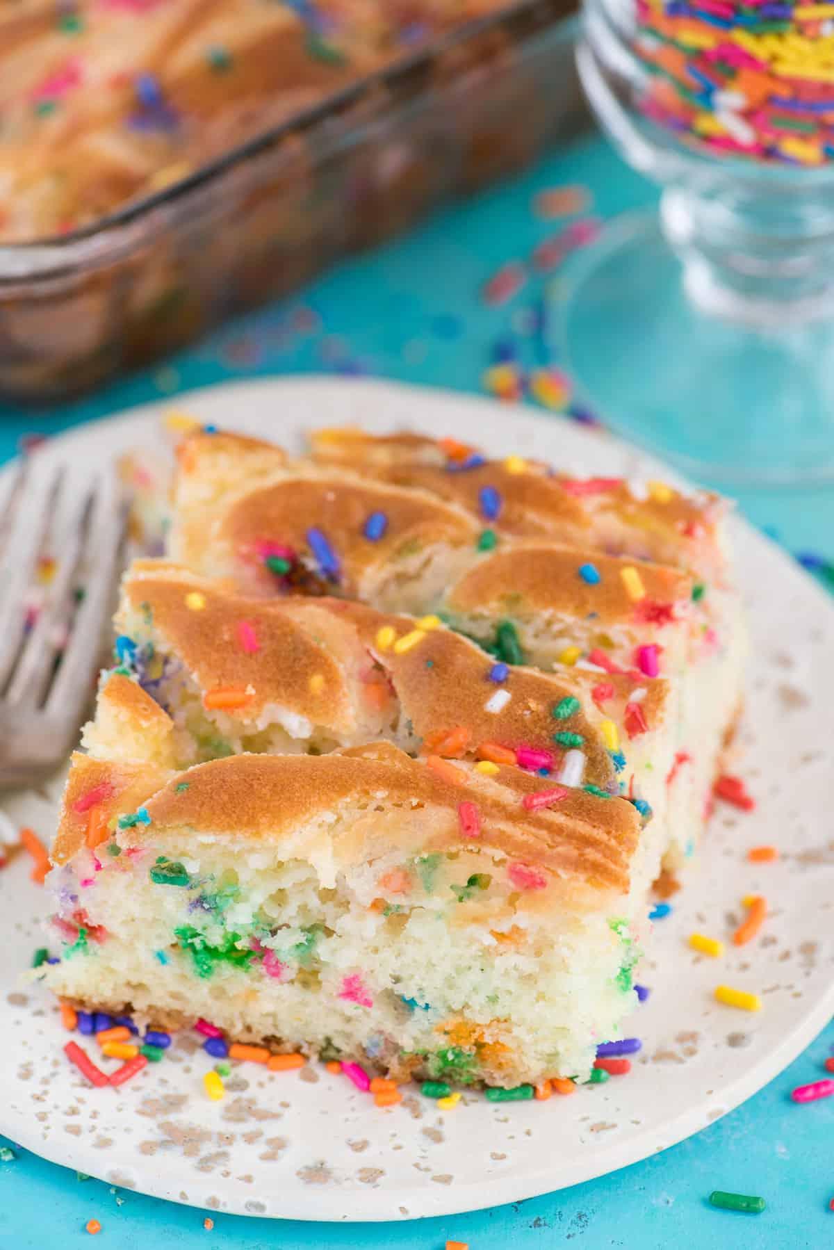 Questa torta terremoto funfetti è un mix per torta bianca arricchito con zuccherini e gocce di cioccolato funfetti. Lo strato di glassa al formaggio cremoso si fonde nella torta creando crepe e fessure in superficie. #earthquakecake #funfetticake #cake Fetta di torta terremoto funfetti su piatto bianco su sfondo blu