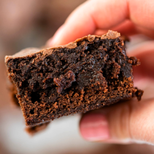 Primo piano di un brownie al cioccolato fudgy tenuto in mano, con la sua crosticina lucida e interna umida.