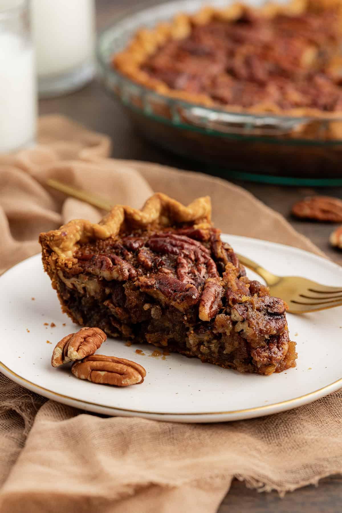 Torta di Noci Pecan Casereccia All'Antica 2 Una fetta di torta di noci pecan su un piatto bianco con una forchetta e noci pecan intere ai lati, poggiata su un telo color tortora su una superficie di legno, con la teglia di vetro piena di torta sullo sfondo e altre noci pecan sparse