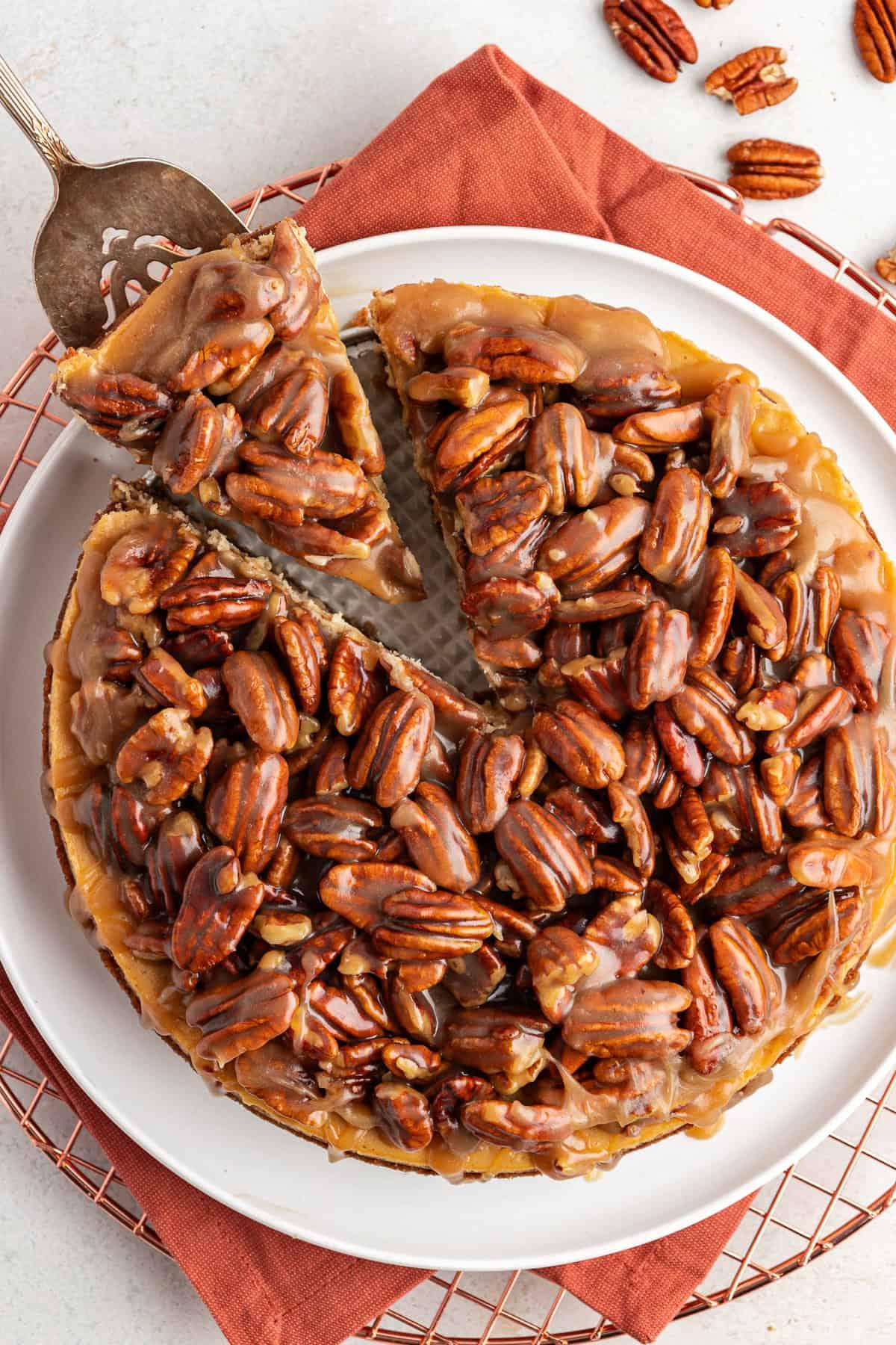 La Migliore Ricetta per Cheesecake alla Torta Pecan 2 Vista dall'alto di una cheesecake alle noci pecan su un panno arancione bruciato su una griglia, con una fetta rimossa da una spatola da portata e noci pecan sparse intorno