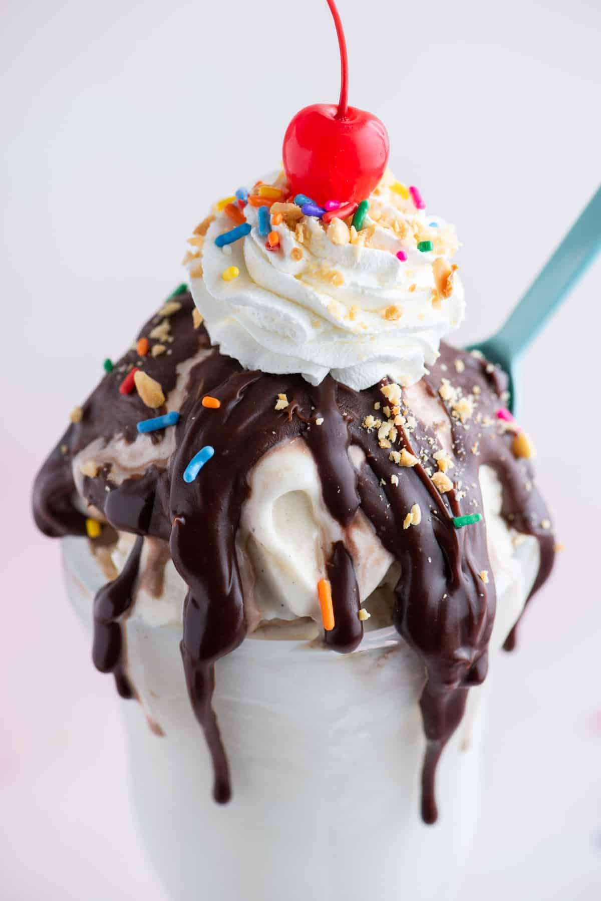 Hot Fudge Sundae 2 Primo piano di un sundae al cioccolato fuso guarnito con codette, arachidi, panna montata e una ciliegia, con un cucchiaio blu infilato nel lato