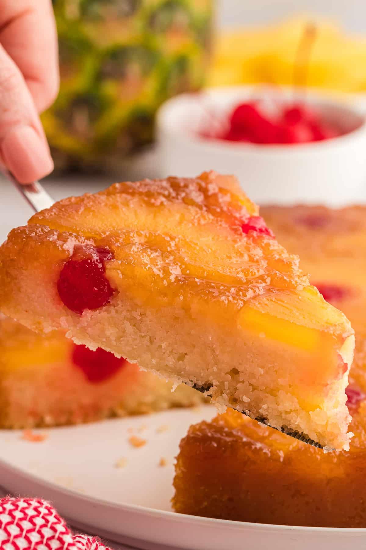 Torta di Ananas Rovesciata Fatta in Casa 2 Una fetta di torta rovesciata all'ananas viene sollevata da un piatto, con un ananas intero, ciliegie e fette di ananas nello sfondo e un canovaccio rosso e bianco accanto al piatto.