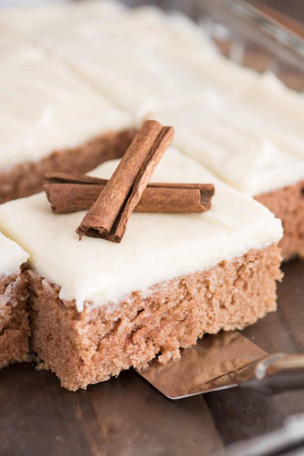 Questa torta speziata alla cannella è una ricetta autunnale incredibile! Conosciuta anche come 'wacky cake', è realizzata senza uova, latte o burro. È la torta autunnale preferita dalla nostra famiglia perché è così facile da fare! Una fetta di torta speziata alla cannella con glassa al formaggio cremoso, guarnita con due stecche di cannella.
