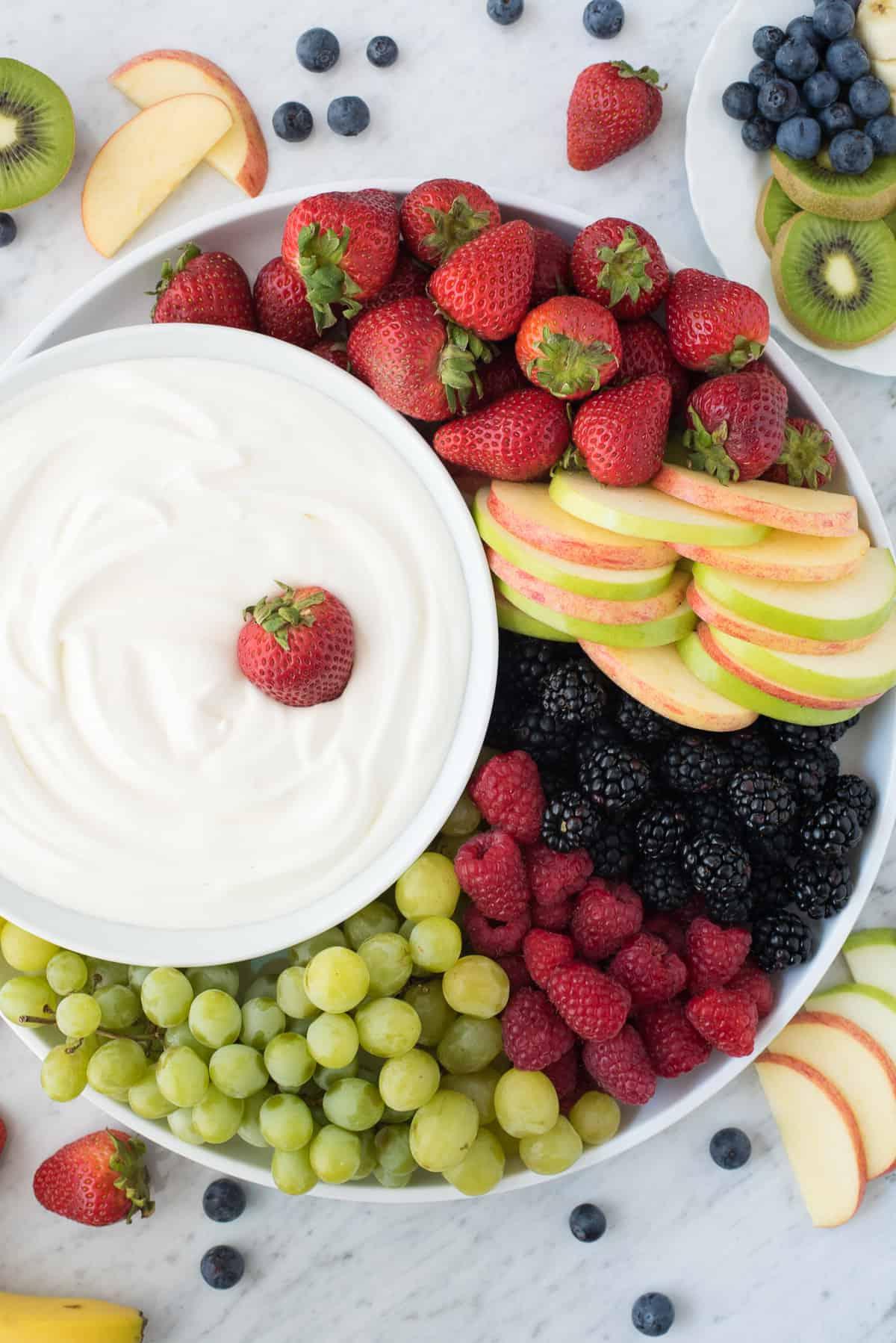 Dip alla frutta con 3 ingredienti! Amiamo questa crema vellutata perché NON c'è formaggio spalmabile! Piatto rotondo pieno di fragole, mele, more, lamponi e uva con una ciotola di dip bianco per frutta al centro