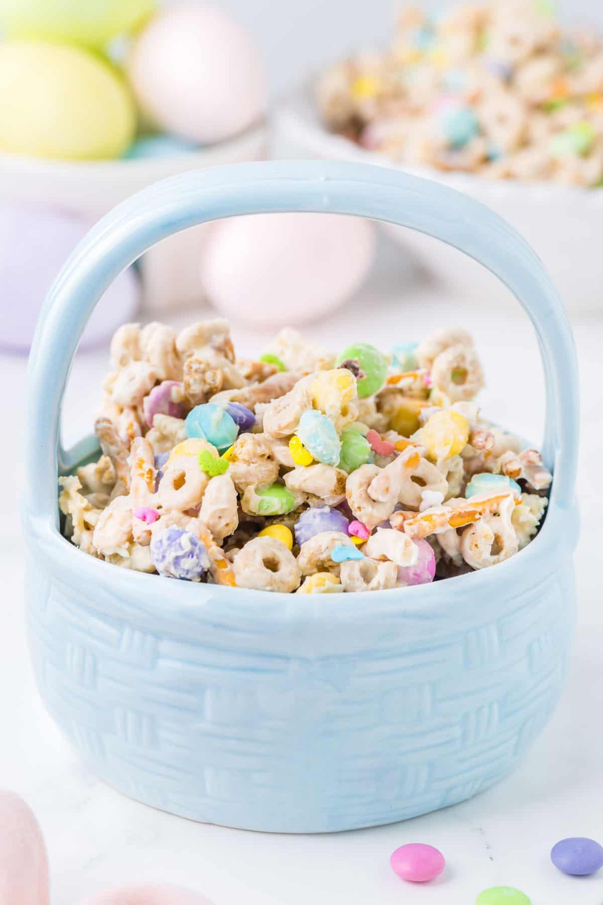 Esca per conigli 2 Snack mix 'Bunny Bait' pasquale in una ciotola blu a forma di cestino pasquale con uova di Pasqua e altro 'Bunny Bait' sullo sfondo e alcune M&M's intorno