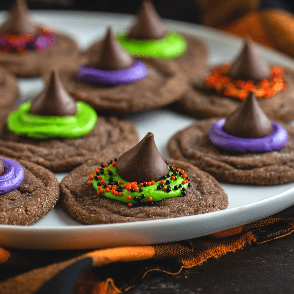 Morbidi biscotti al cioccolato sormontati da un bacio di cioccolato, glassa e zuccherini, questi Biscotti Cappello da Strega sono un dessert di Halloween facile. Un piatto bianco con biscotti a forma di cappello da strega