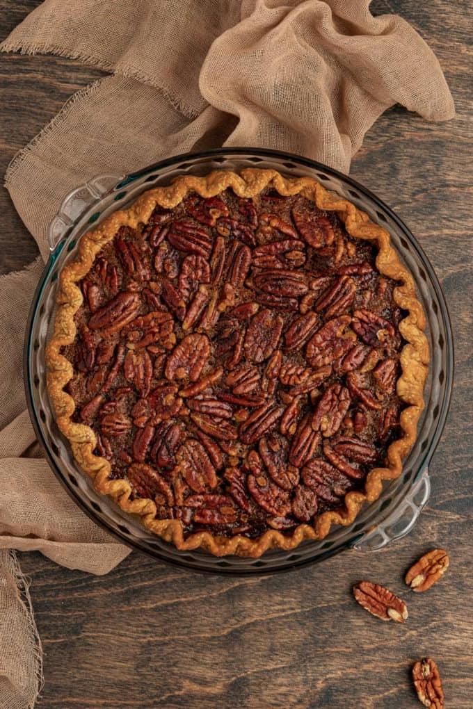 Torta di Noci Pecan Casereccia All'Antica 11 Una torta di noci pecan appena sfornata in una teglia di vetro, poggiata su una superficie di legno con un telo color tortora e noci pecan intere intorno