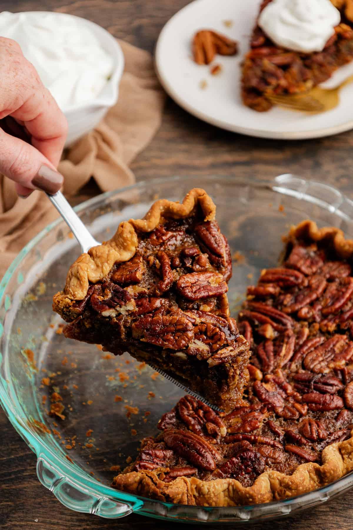 Torta di Noci Pecan Casereccia All'Antica 12 Una fetta di torta di noci pecan viene sollevata da una teglia di vetro semi-vuota poggiata su una superficie di legno accanto a un telo color tortora, un piatto con una fetta di torta guarnita con panna montata, e una ciotola di panna montata.