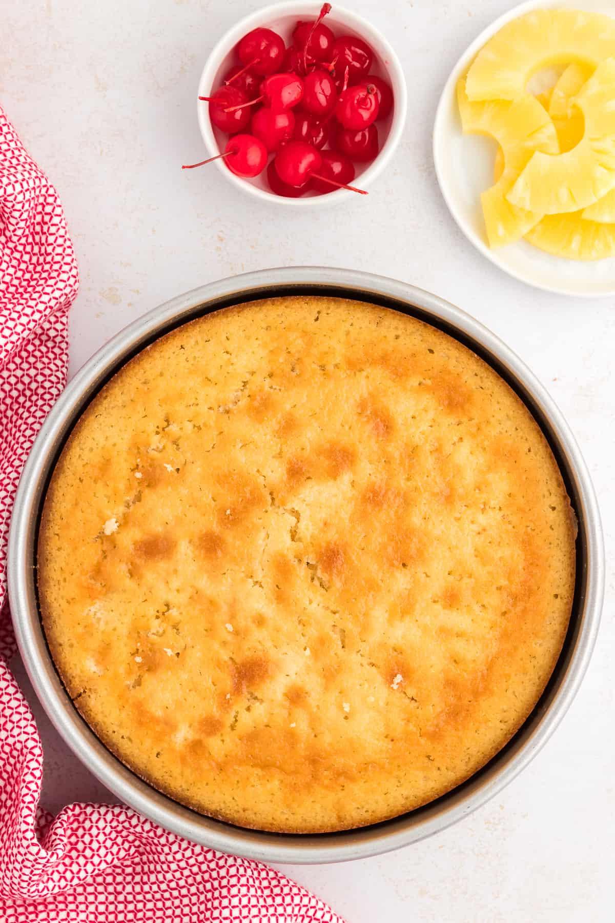 Torta di Ananas Rovesciata Fatta in Casa 12 Torta rovesciata all'ananas appena sfornata, prima di essere capovolta, accanto a un piatto di fette di ananas e una ciotola di ciliegie al maraschino e un canovaccio rosso e bianco.