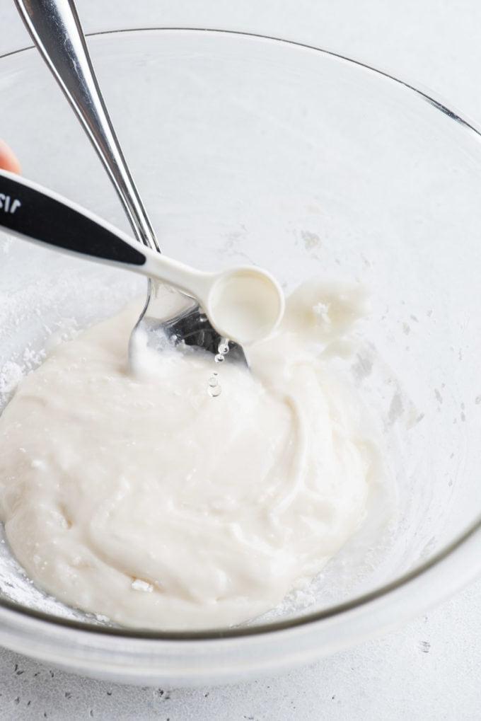 Glassa Reale senza Polvere di Meringa 13 ghiaccia reale in una ciotola di vetro trasparente con un cucchiaio e gocce d'acqua aggiunte con un piccolo cucchiaino dosatore