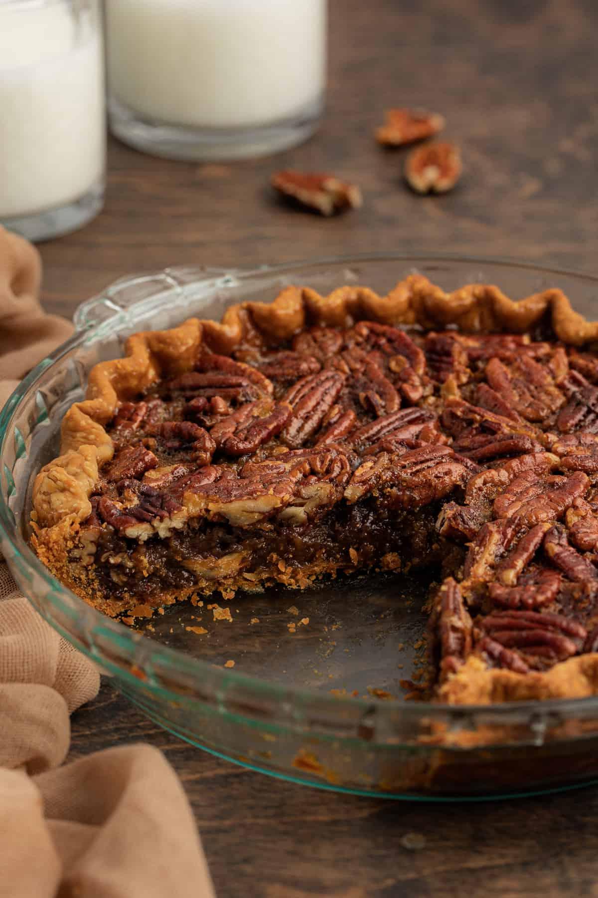 Torta di Noci Pecan Casereccia All'Antica 13 Torta di noci pecan con una fetta grande mancante in una teglia di vetro, poggiata su una superficie di legno circondata da noci pecan e un telo color tortora con due bicchieri di latte sullo sfondo