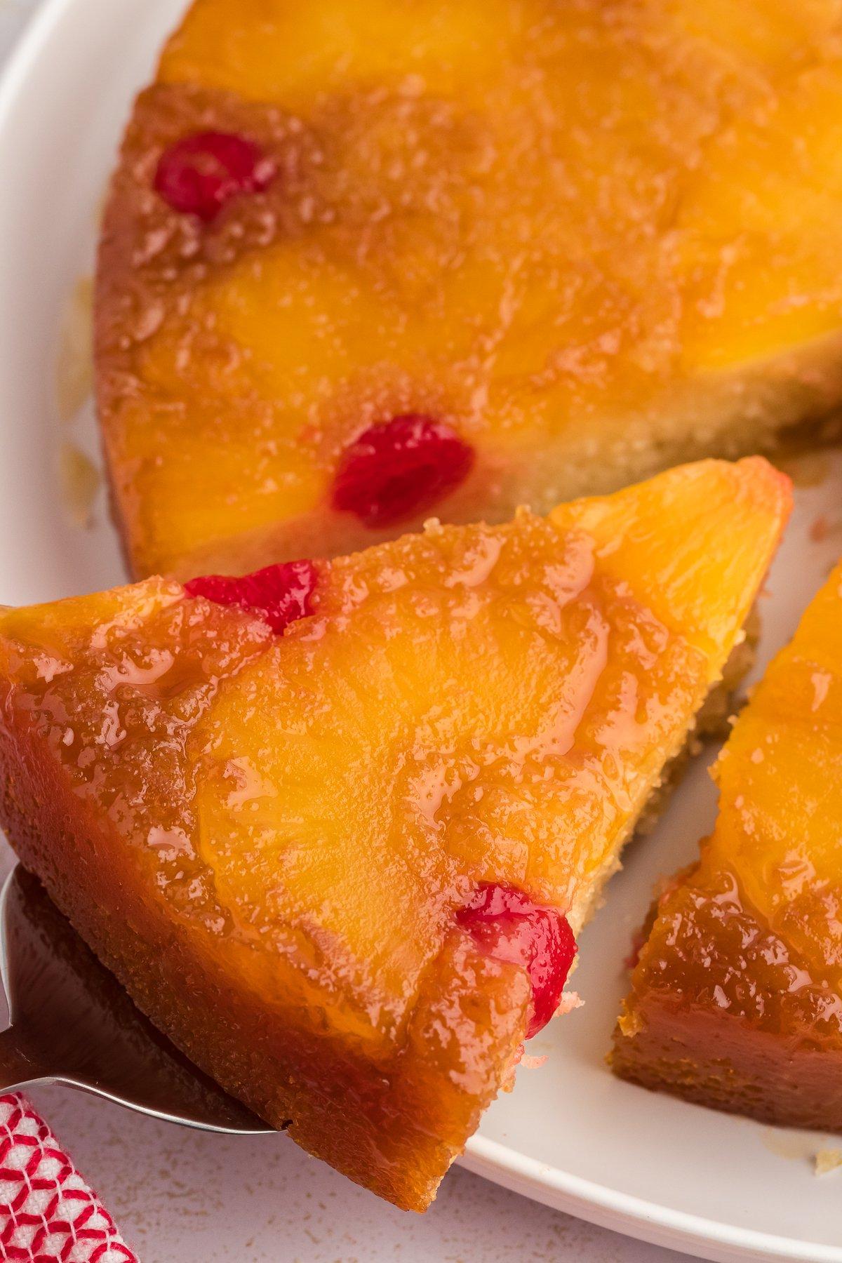 Torta di Ananas Rovesciata Fatta in Casa 13 Primo piano di una fetta di torta rovesciata all'ananas che viene sollevata dal piatto, lontano dal resto della torta.