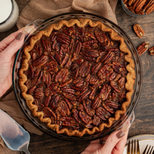 Torta di Noci Pecan Casereccia All'Antica 14 Una torta di noci pecan in una teglia di vetro su una superficie di legno con un telo color tortora, noci pecan, una spatola per servire, piatti e forchette intorno
