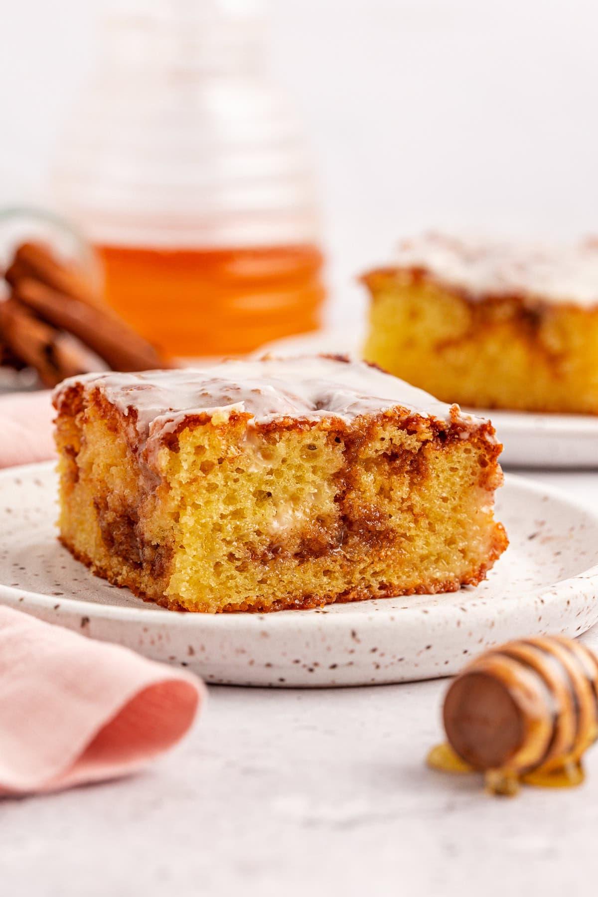 Torta Girella al Miele 14 Primo piano di una fetta di torta di girelle alla cannella con glassa su un piatto maculato. Sullo sfondo, un'altra fetta di torta Honey Bun è su un piatto, e un mestolo per miele con gocce di miele è visibile. Un panno rosa chiaro è parzialmente mostrato.