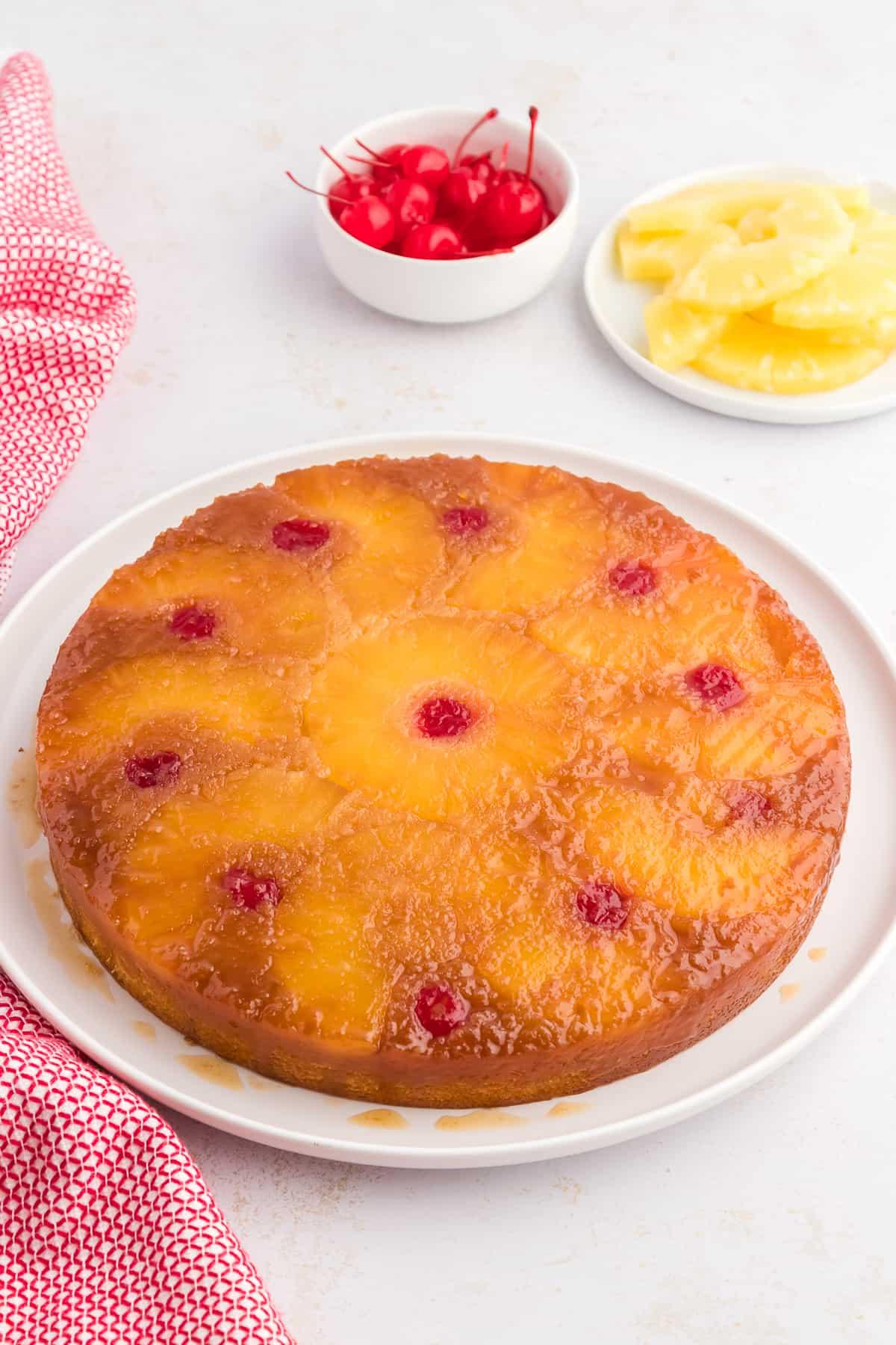Torta di Ananas Rovesciata Fatta in Casa 14 Torta rovesciata all'ananas su un piatto bianco circondata da fette di ananas, ciliegie al maraschino e un canovaccio rosso e bianco.