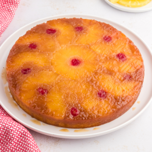 Torta di Ananas Rovesciata Fatta in Casa 15 Torta rovesciata all'ananas su un piatto bianco circondata da fette di ananas, ciliegie al maraschino e un canovaccio rosso e bianco.