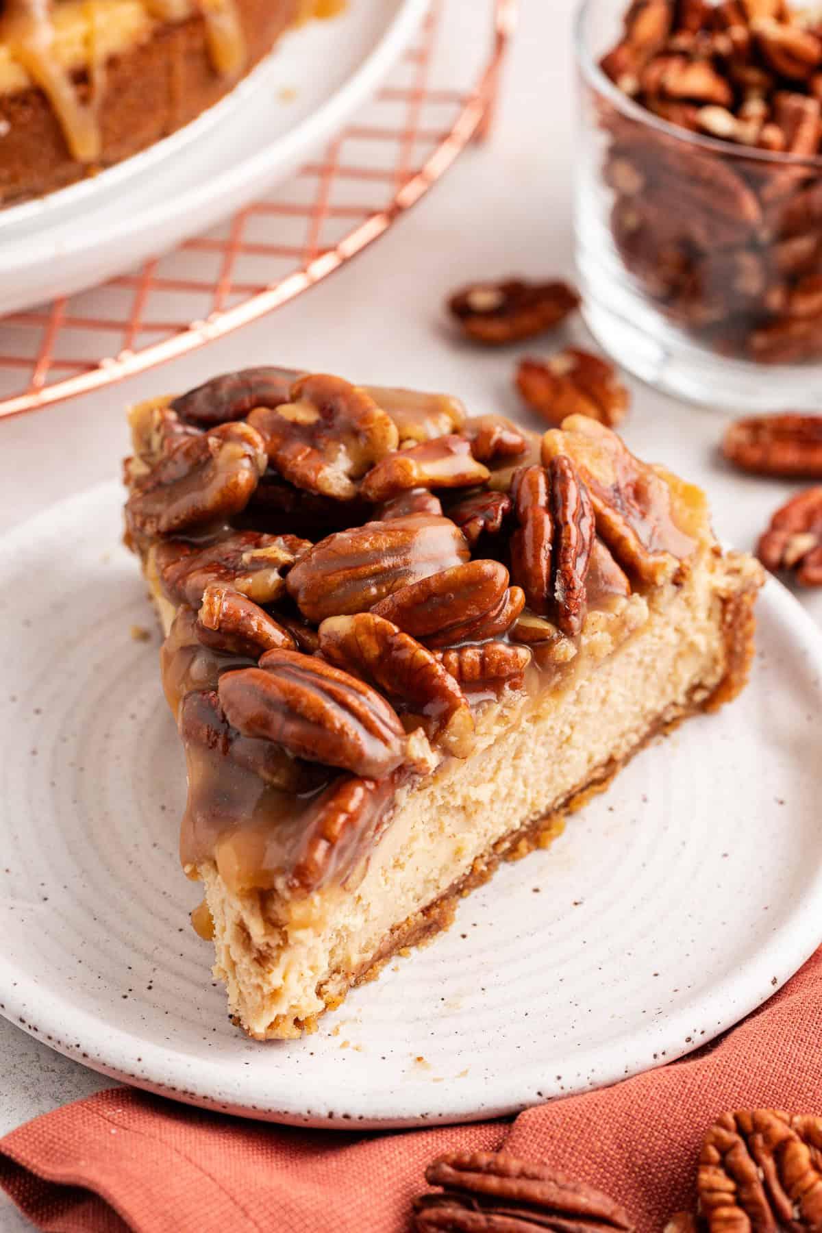 La Migliore Ricetta per Cheesecake alla Torta Pecan 16 Una fetta di cheesecake alle noci pecan su un piattino bianco accanto a un panno arancione bruciato con noci pecan sparse intorno