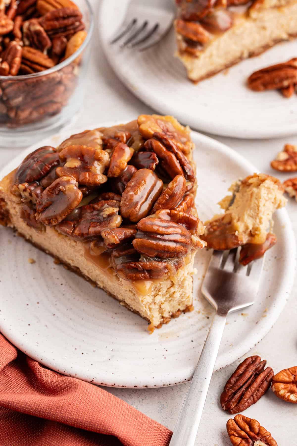 La Migliore Ricetta per Cheesecake alla Torta Pecan 17 Una fetta di cheesecake alle noci pecan su un piattino bianco con una forchetta accanto e un boccone di cheesecake su di essa, circondata da un panno arancione bruciato, noci pecan sparse, una ciotola di vetro di noci pecan e un'altra fetta su un piatto separato
