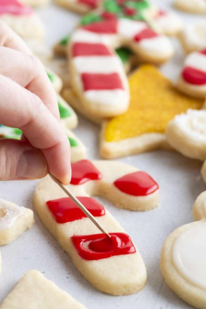 Glassa Reale senza Polvere di Meringa 18 ghiaccia reale rossa spalmata con uno stuzzicadenti su un biscotto a forma di bastoncino di zucchero con altri biscotti a tema natalizio sullo sfondo