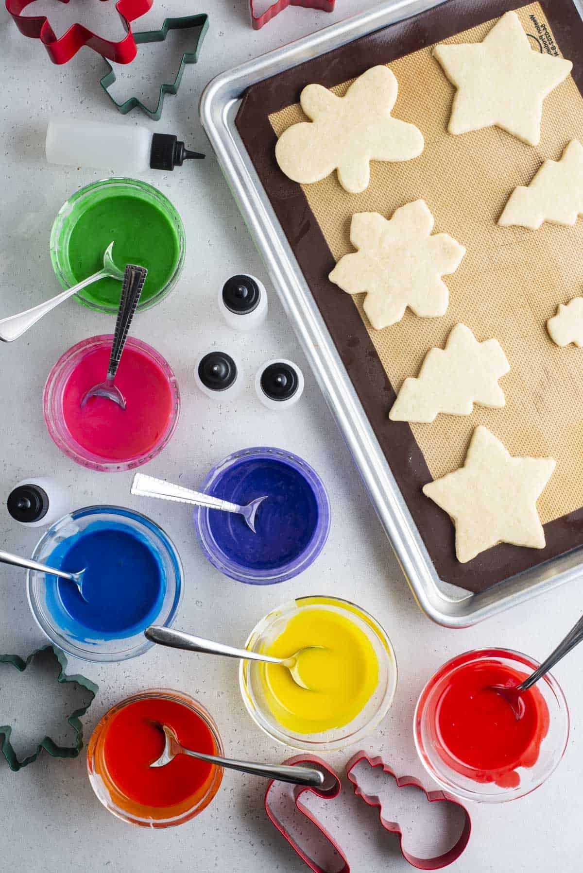 Glassa Reale senza Polvere di Meringa 20 biscotti a forma di Natale allineati in file su una teglia da forno con ciotole di glassa colorata con cucchiai accanto, bottiglie a spremere vuote e formine per biscotti