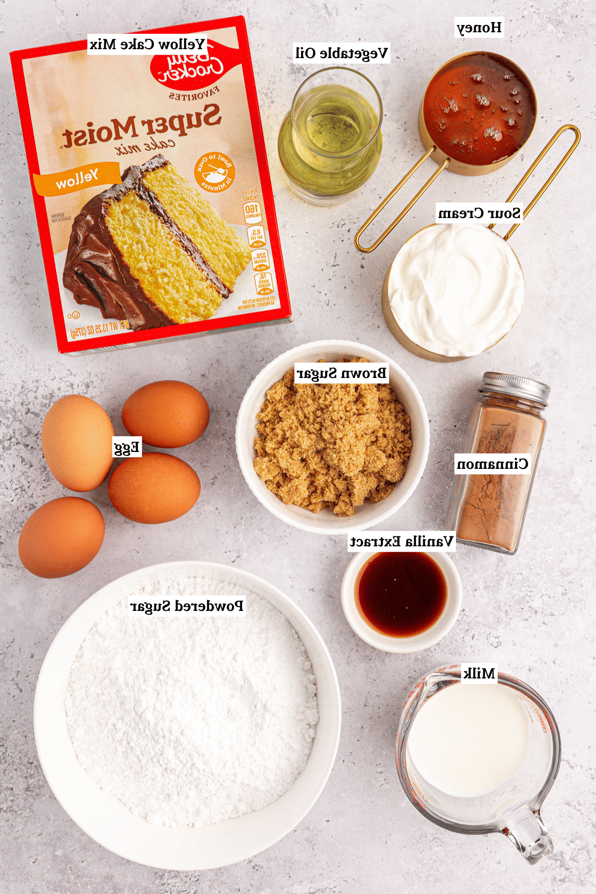 Torta Girella al Miele 3 Vista dall'alto degli ingredienti per la torta Honey Bun disposti su una superficie chiara, con una scatola di preparato per torte giallo, uova, panna acida, latte, miele, olio vegetale, zucchero di canna, cannella, estratto di vaniglia e zucchero a velo.