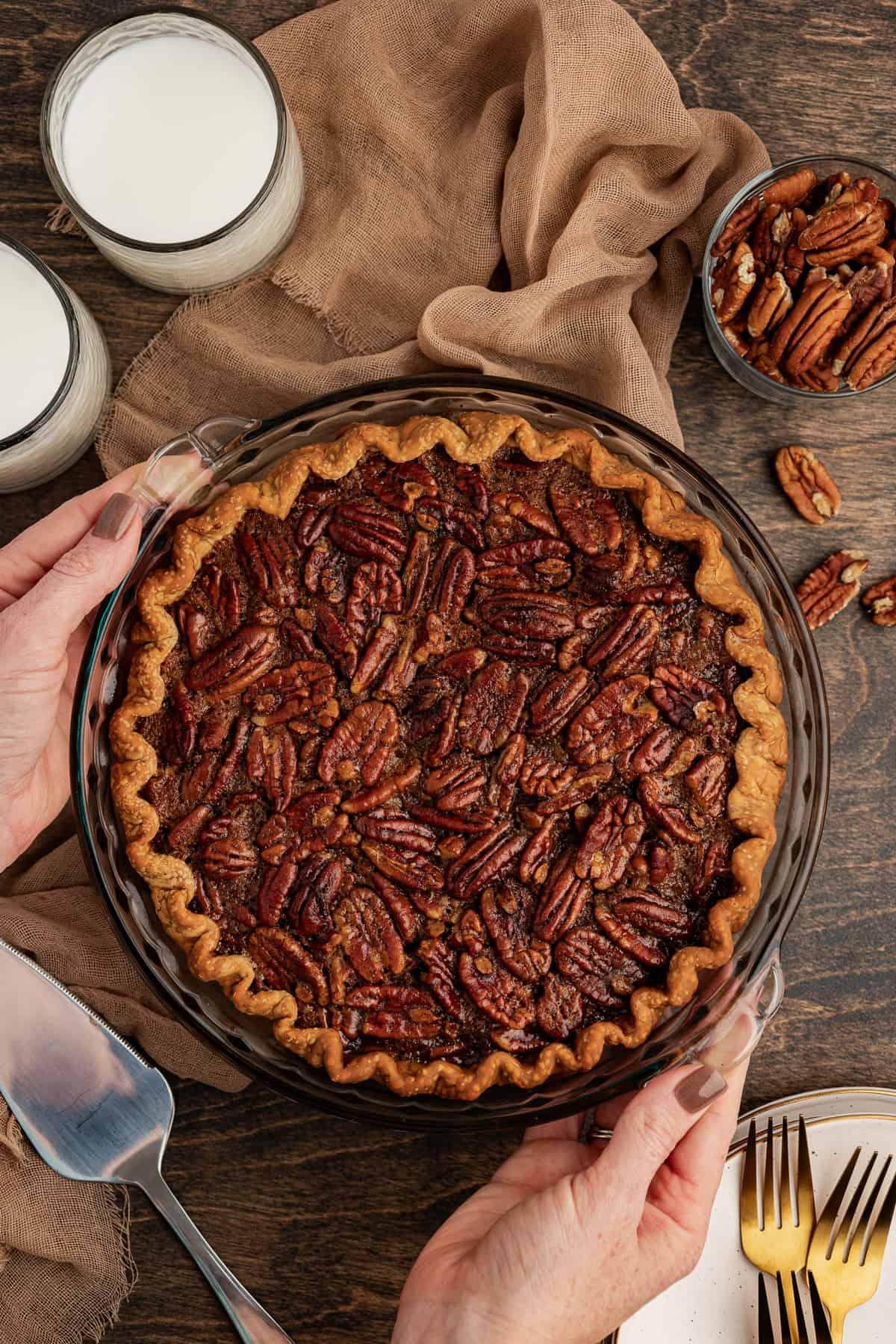 Torta di Noci Pecan Casereccia All'Antica 3 Una torta di noci pecan intera viene appoggiata su una superficie di legno con due mani, su un telo color tortora e circondata da due bicchieri di latte, una piccola ciotola di noci pecan, altre noci pecan sparse, una pila di piatti e forchette e una spatola per servire la torta