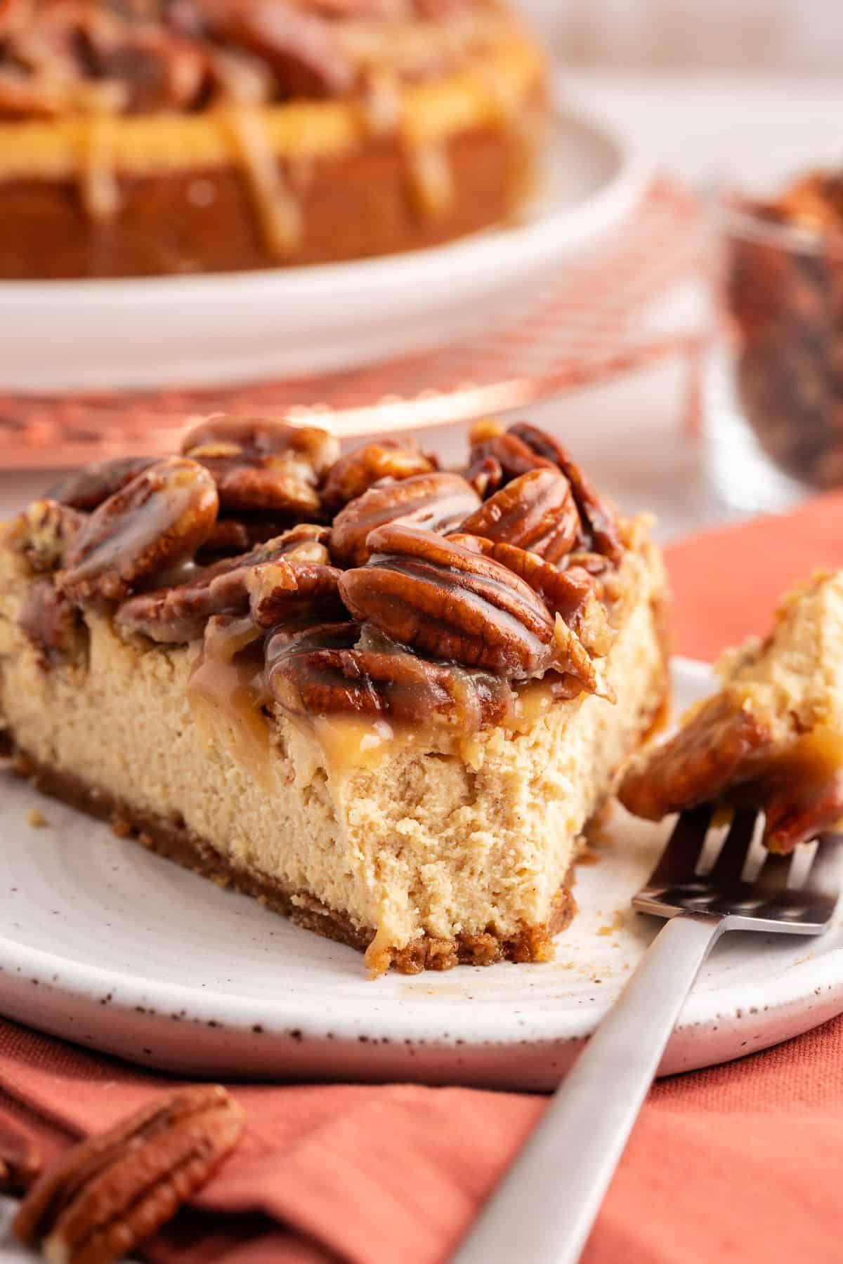 La Migliore Ricetta per Cheesecake alla Torta Pecan 3 Una fetta di cheesecake alle noci pecan su un piattino bianco con una forchetta con un boccone sulla parte laterale del piatto, su un panno arancione bruciato con noci pecan sparse e l'intera torta sullo sfondo