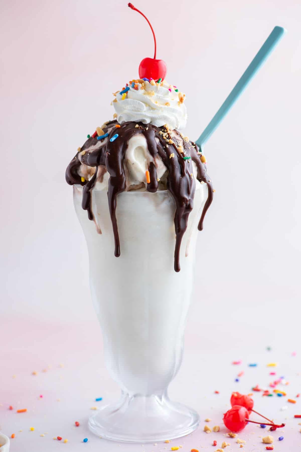 Hot Fudge Sundae 3 Un sundae al cioccolato fuso in un bicchiere alto circondato da codette, pezzi di arachidi e un paio di ciliegie al maraschino, con un cucchiaio blu infilato nel lato del sundae