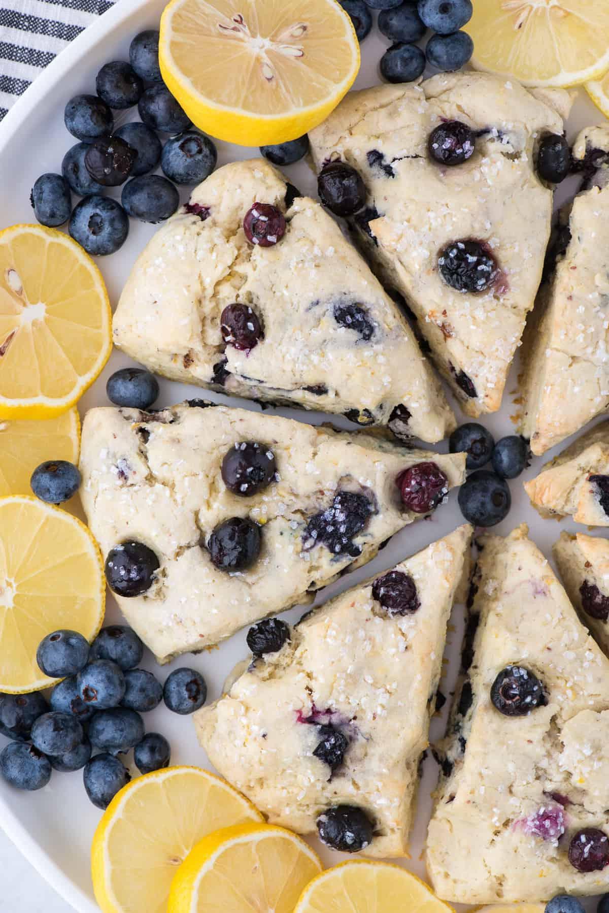 I Migliori Scones al Limone e Mirtilli 3 Scones al limone e mirtilli disposti in cerchio su un piatto bianco circondati da fette di limone e mirtilli