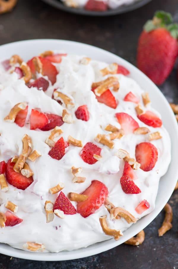 Primo piano di una ciotola di Insalata Dolce Fragole e Pretzel, con i suoi strati invitanti di crema morbida, pezzi di fragola rossa brillante e la croccantezza dei pretzel tritati, una vera delizia estiva.