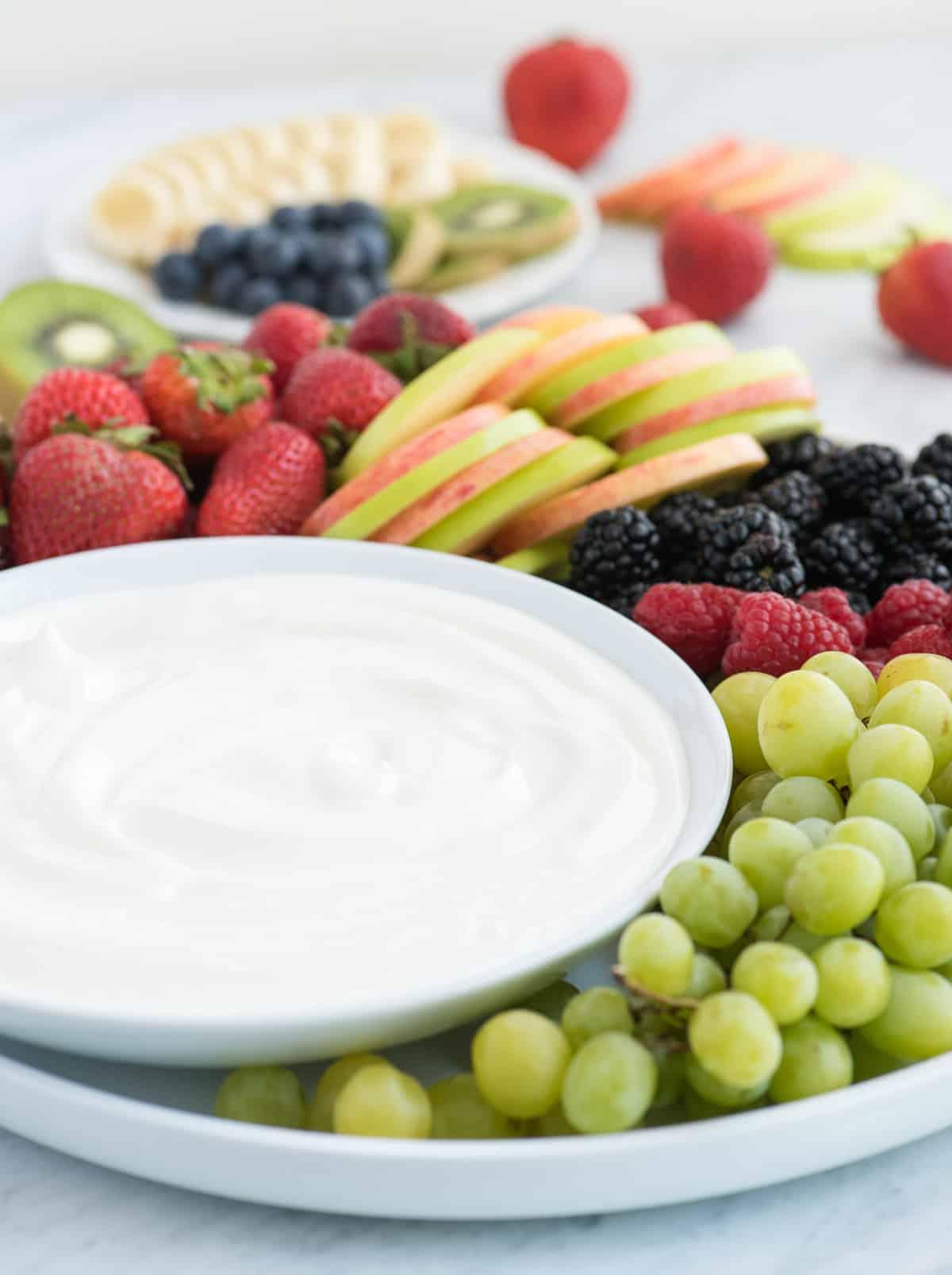 Dip alla frutta con 3 ingredienti! Amiamo questa crema vellutata perché NON c'è formaggio spalmabile! Piatto rotondo pieno di fragole, mele, more, lamponi e uva con una ciotola di dip bianco per frutta al centro