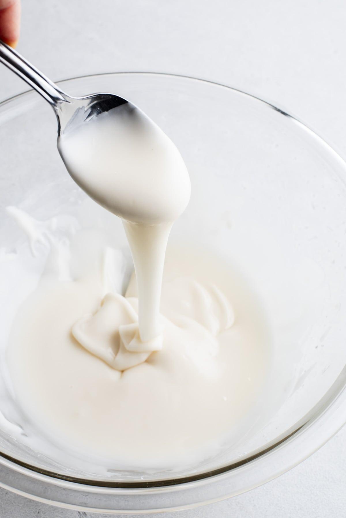Glassa Reale senza Polvere di Meringa 3 ghiaccia reale in una ciotola di vetro trasparente con un cucchiaio pieno di glassa sopra e glassa che cola nella ciotola dal cucchiaio