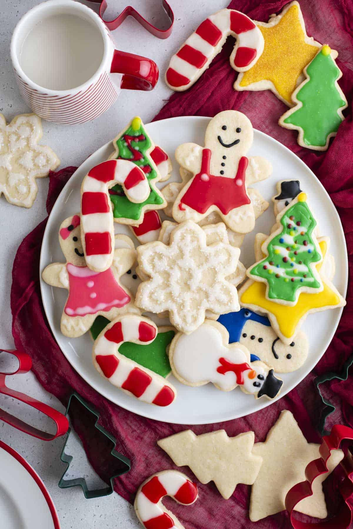 Glassa Reale senza Polvere di Meringa 22 un piatto pieno di biscotti natalizi decorati con ghiaccia reale su un asciugamano rosso con altri biscotti, formine per biscotti e una tazza di latte intorno