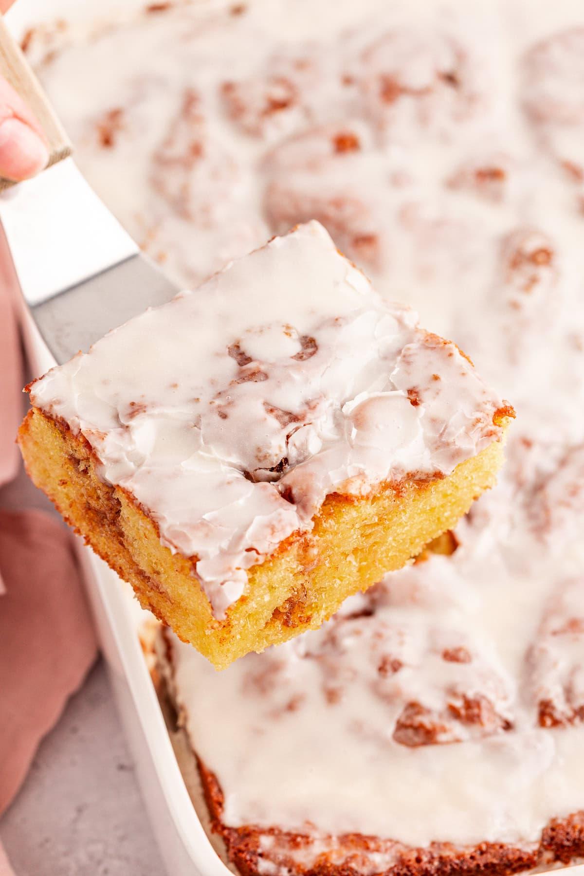 Torta Girella al Miele 4 Primo piano di una fetta di torta Honey Bun glassata, sollevata da un vassoio con una spatola. La torta è dorata con una glassa bianca e visibili spirali di cannella.