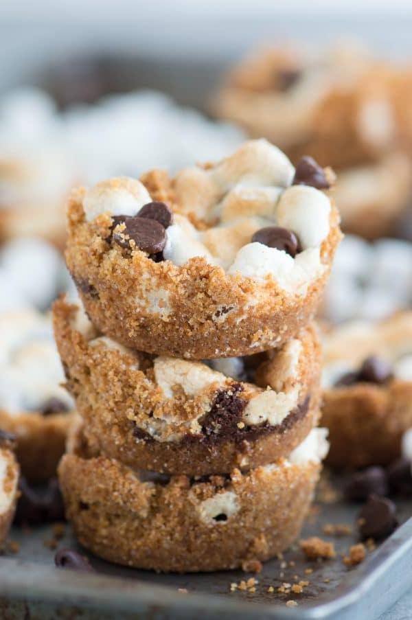 Tazze S'mores al Forno con 5 ingredienti! Preparale in uno stampo per muffin standard. Un incrocio tra biscotti s'mores e cupcake s'mores! Tre Tazze S'mores facili impilate, circondate da altre tazze s'mores fatte in casa su una teglia.