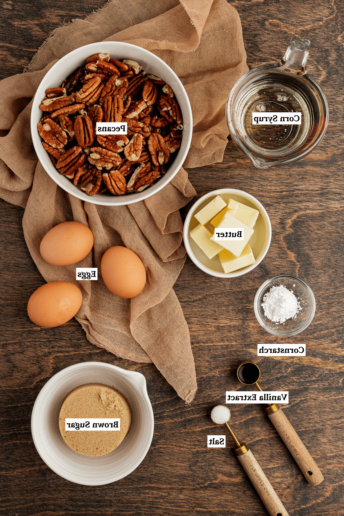 Torta di Noci Pecan Casereccia All'Antica 4 Ingredienti per la torta di noci pecan disposti su una superficie di legno con un telo color tortora, inclusi sciroppo di mais, noci pecan, burro, uova, amido di mais, estratto di vaniglia, sale e zucchero di canna