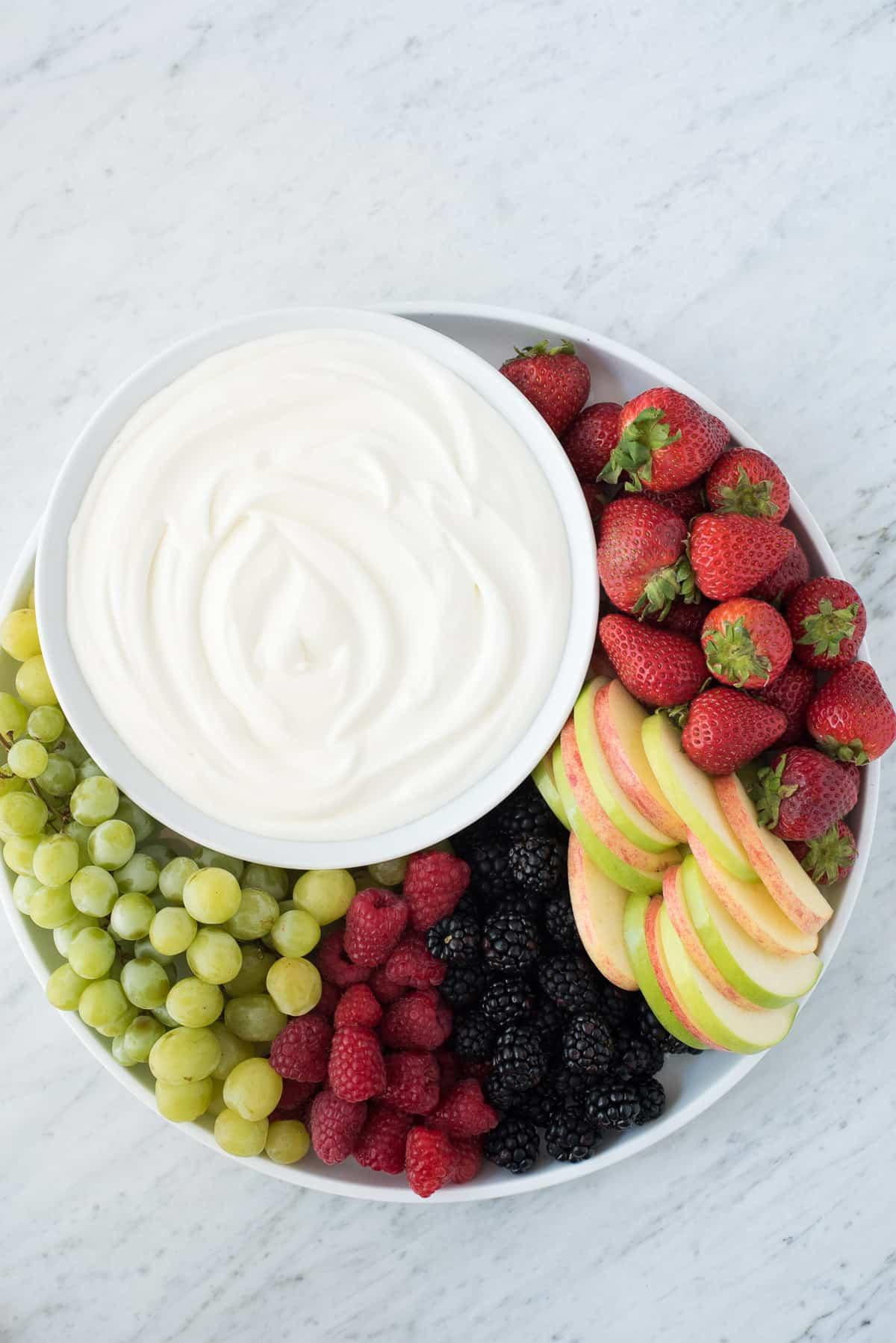 Dip alla frutta con 3 ingredienti! Amiamo questa crema vellutata perché NON c'è formaggio spalmabile! Piatto rotondo pieno di fragole, mele, more, lamponi e uva con una ciotola di dip bianco per frutta al centro