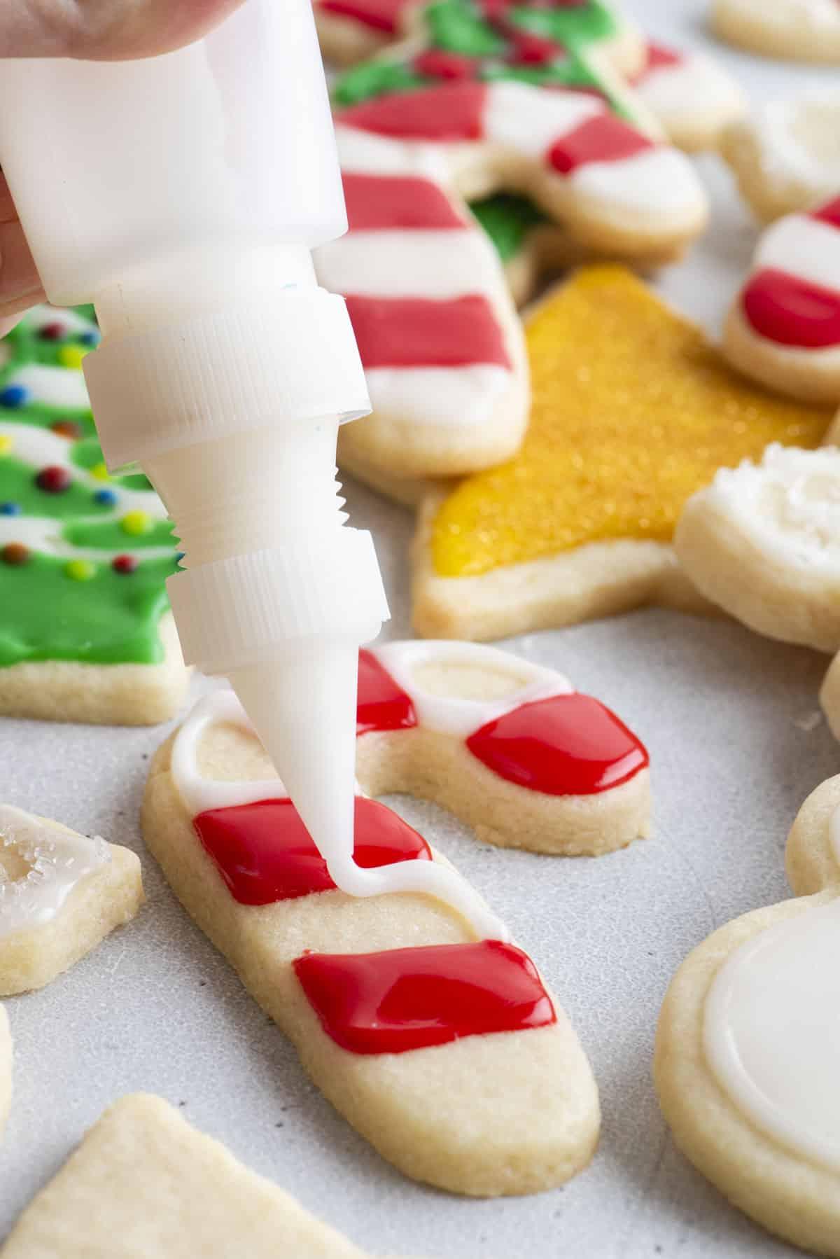 Glassa Reale senza Polvere di Meringa 4 ghiaccia reale bianca che viene applicata tra ghiaccia rossa su un biscotto a forma di bastoncino di zucchero con altri biscotti a tema natalizio sullo sfondo