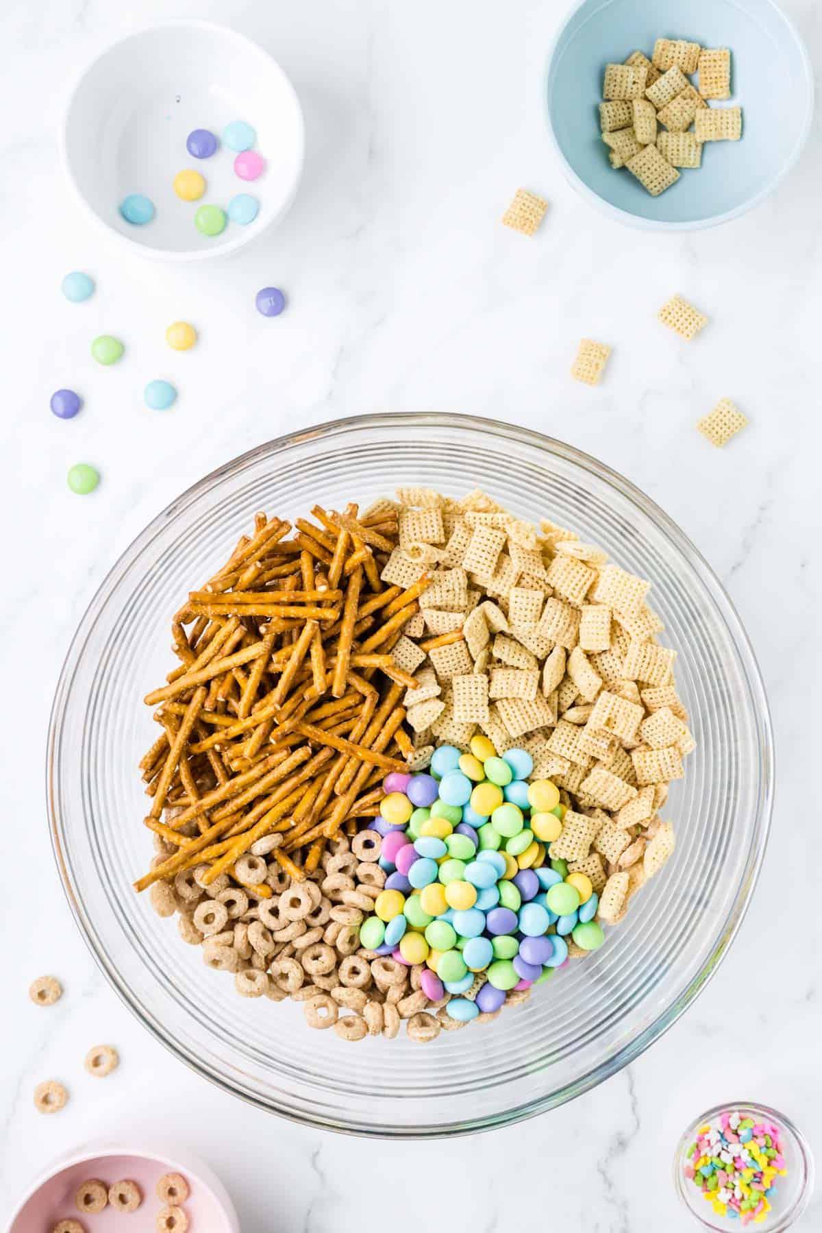 Esca per conigli 5 Ingredienti secchi per 'Bunny Bait' in una ciotola di vetro trasparente circondata da una ciotola di codette, una ciotola con Cheerios, una ciotola di Chex di riso e una ciotola di M&M's