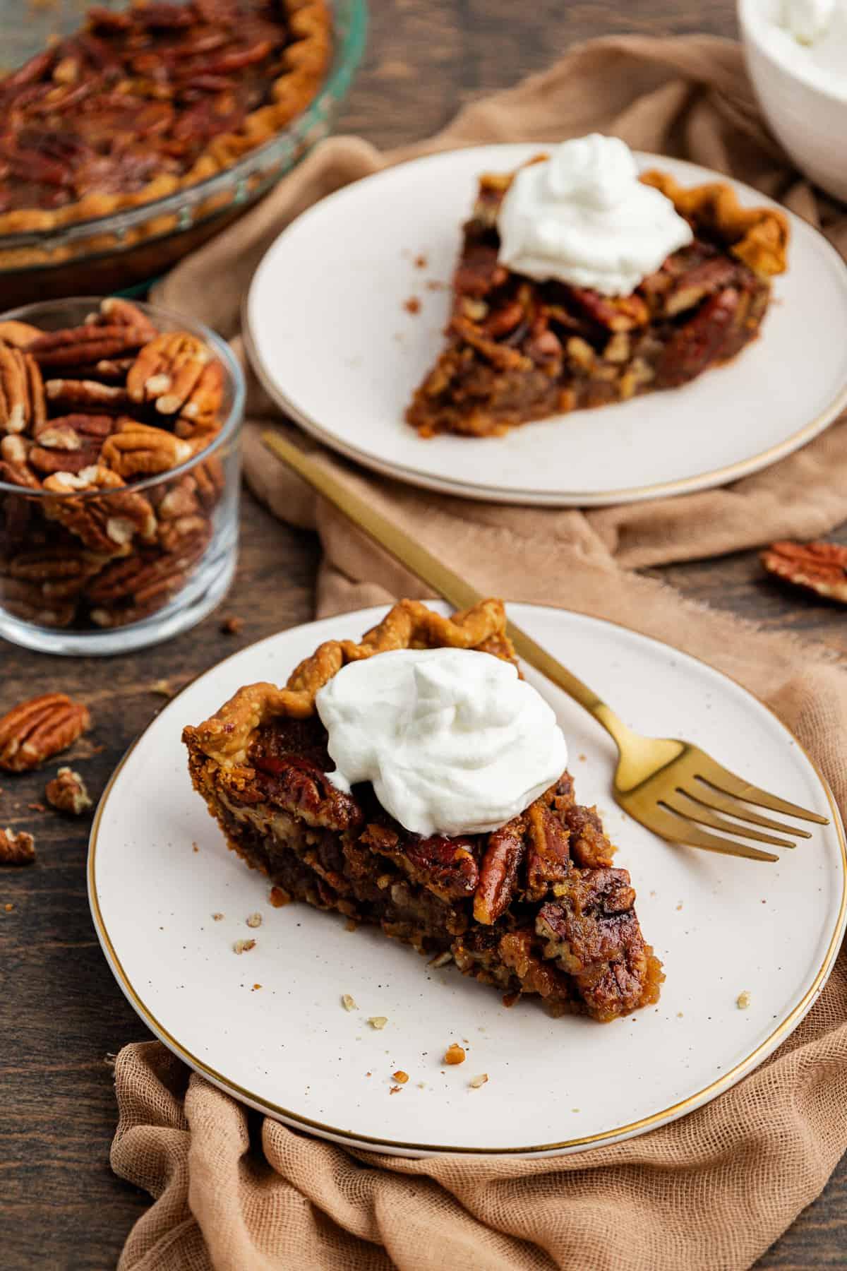 Torta di Noci Pecan Casereccia All'Antica 5 Due piatti bianchi con fette di torta di noci pecan, con una forchetta sul piatto anteriore, poggiati su un telo color tortora su una superficie di legno con una ciotola di vetro di noci pecan accanto e altre noci pecan sparse, e la teglia con altra torta sullo sfondo