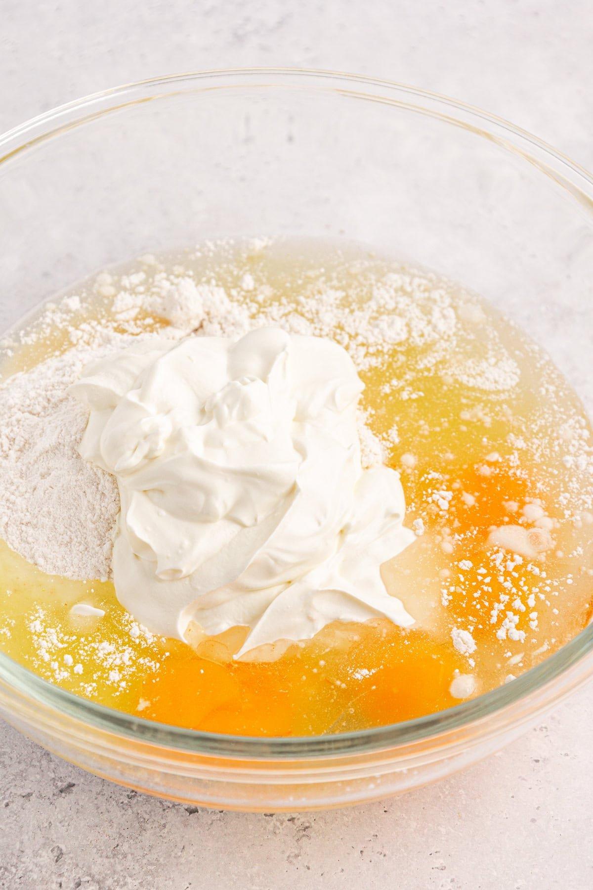 Torta Girella al Miele 5 Una ciotola di vetro con ingredienti per l'impasto della torta Honey Bun contiene due uova rotte, farina, una cucchiaiata di panna acida e un accenno di cannella, pronti per essere mescolati. Lo sfondo è una superficie testurizzata grigio chiaro.