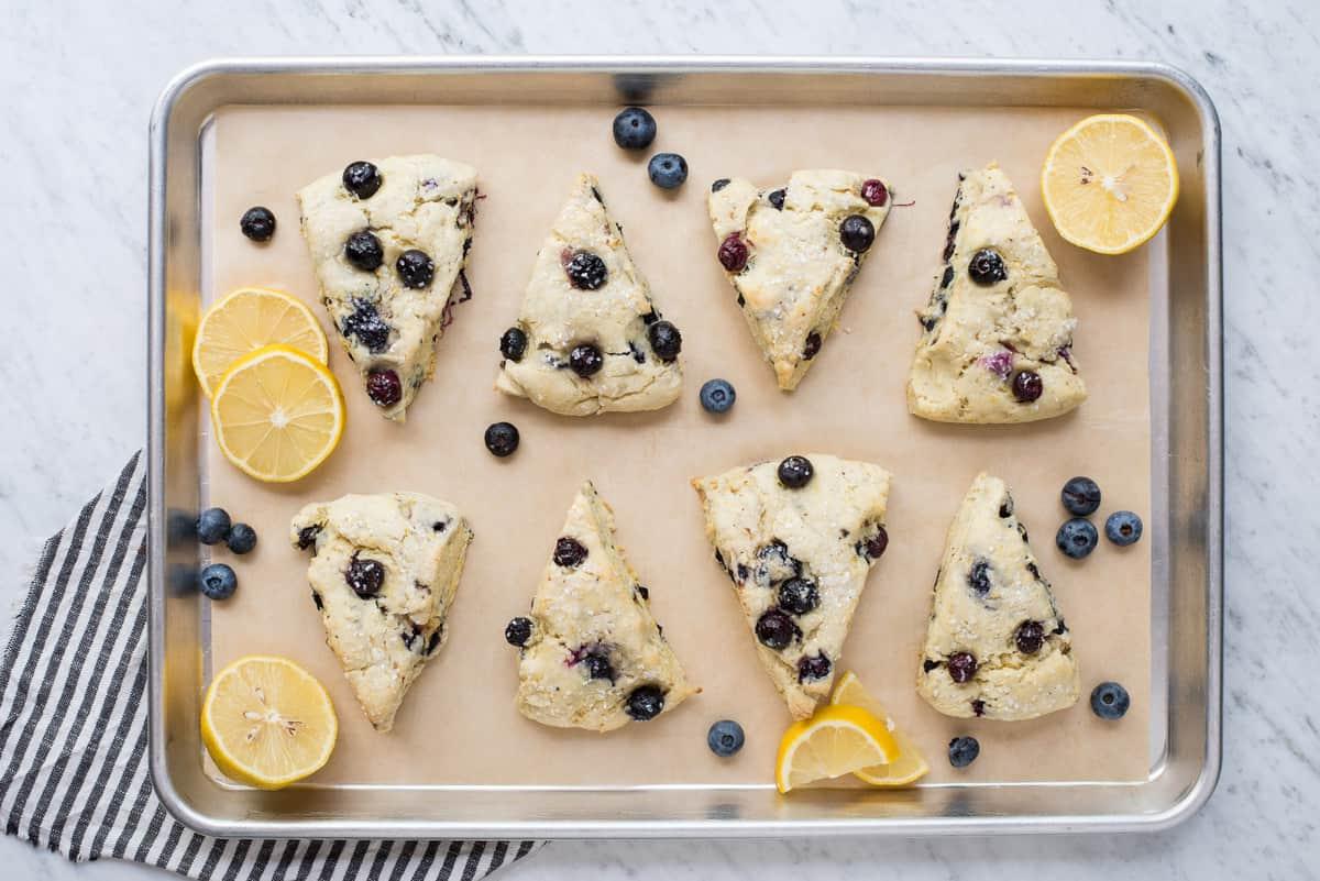 I Migliori Scones al Limone e Mirtilli 5 Scones al limone e mirtilli su carta forno marrone con fette di limone e mirtilli