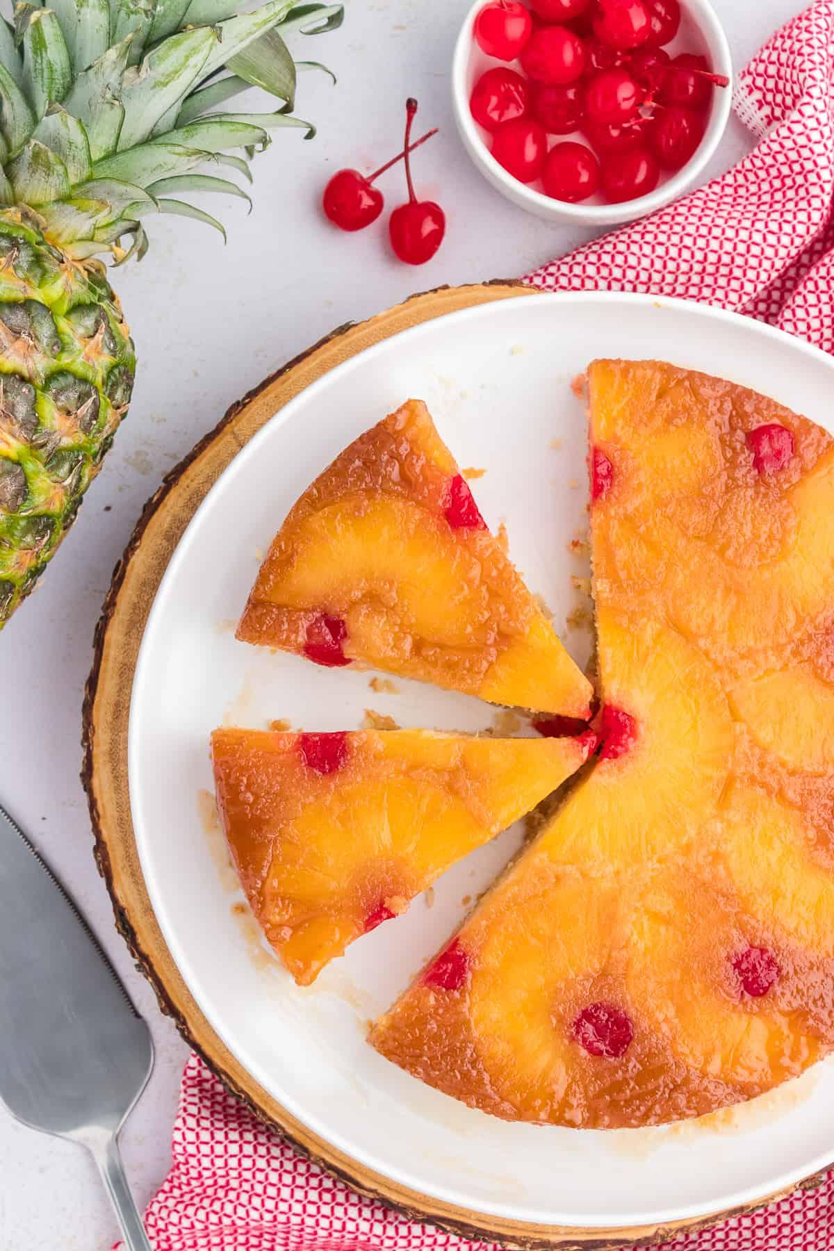 Torta di Ananas Rovesciata Fatta in Casa 5 Fetta di torta rovesciata all'ananas su un piatto bianco con un canovaccio rosso e bianco, una spatola da portata, ananas e una piccola ciotola di ciliegie al maraschino disposti intorno.
