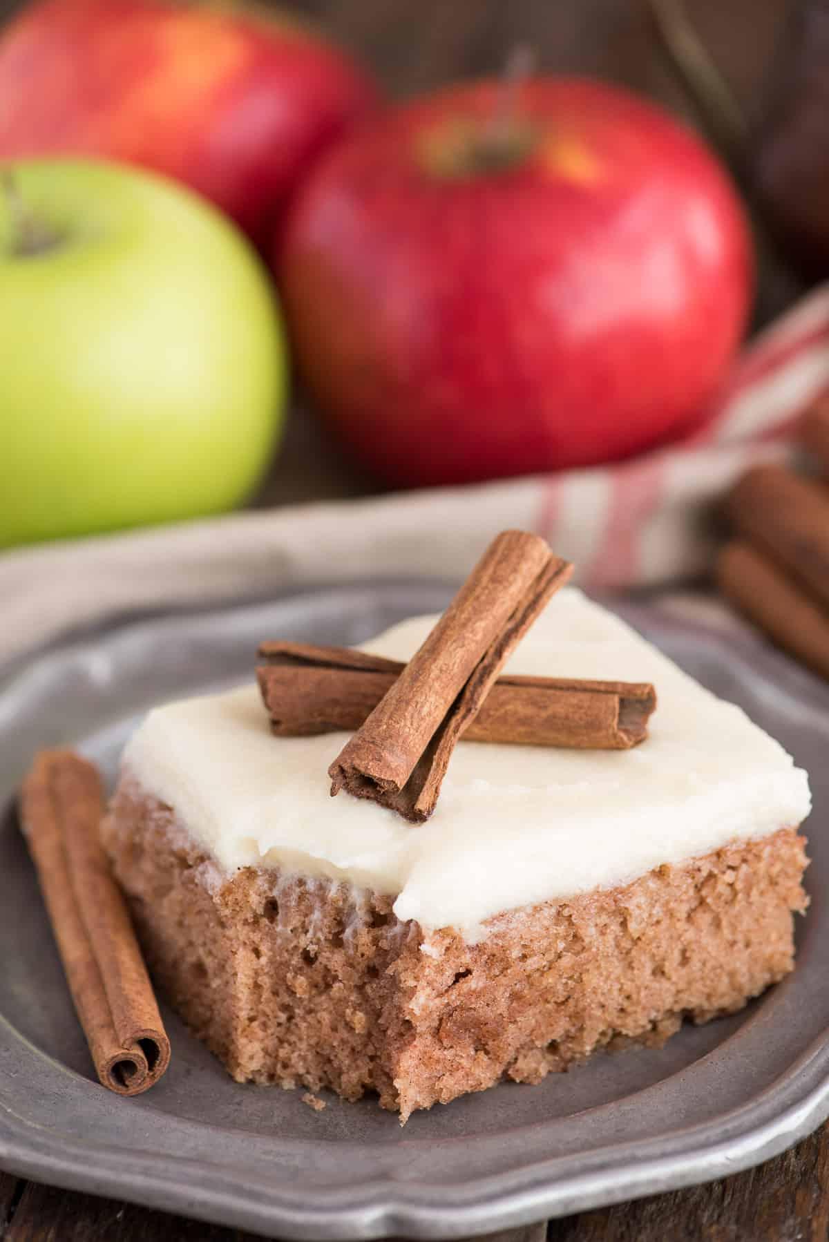 Questa torta speziata alla cannella è una ricetta autunnale incredibile! Conosciuta anche come 'wacky cake', è realizzata senza uova, latte o burro. È la torta autunnale preferita dalla nostra famiglia perché è così facile! Una fetta di torta speziata alla cannella con glassa al formaggio cremoso e due stecche di cannella, su un piatto di metallo.